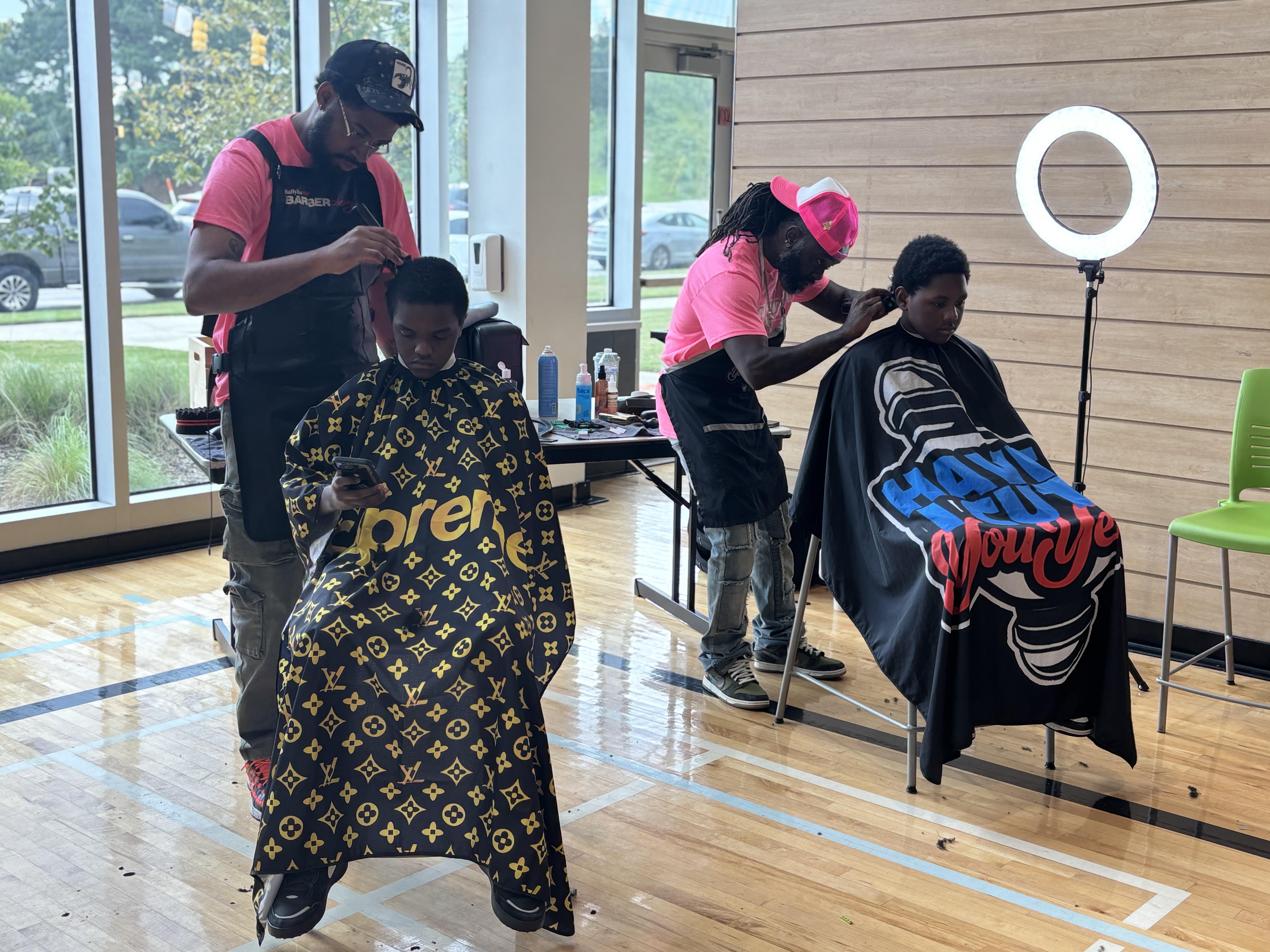 Two barbers in pink foundation shirts give free haircuts to students in the gym at the 2025 Back to School Bash.